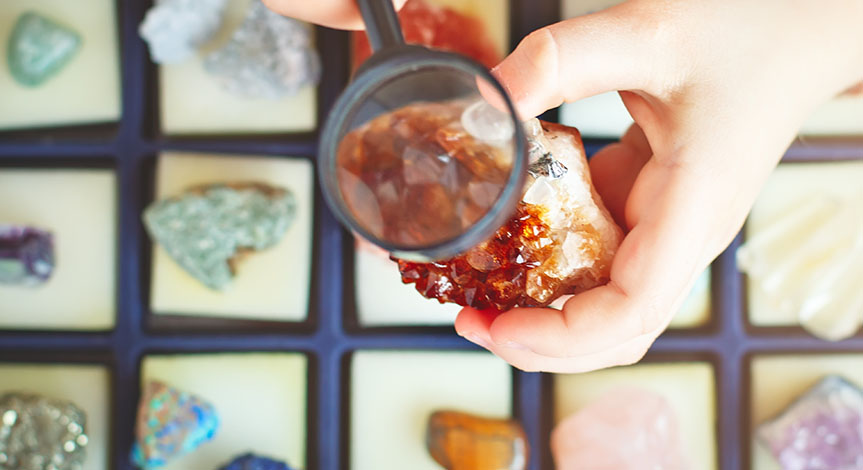 Child holding a mineral in his hand while he looks closely using a magnifying glass over a case full of other minerals