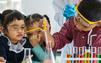 Boy drops something into orange beaker using a pipette in a lab while 3 little girls watch all wearing safety goggles