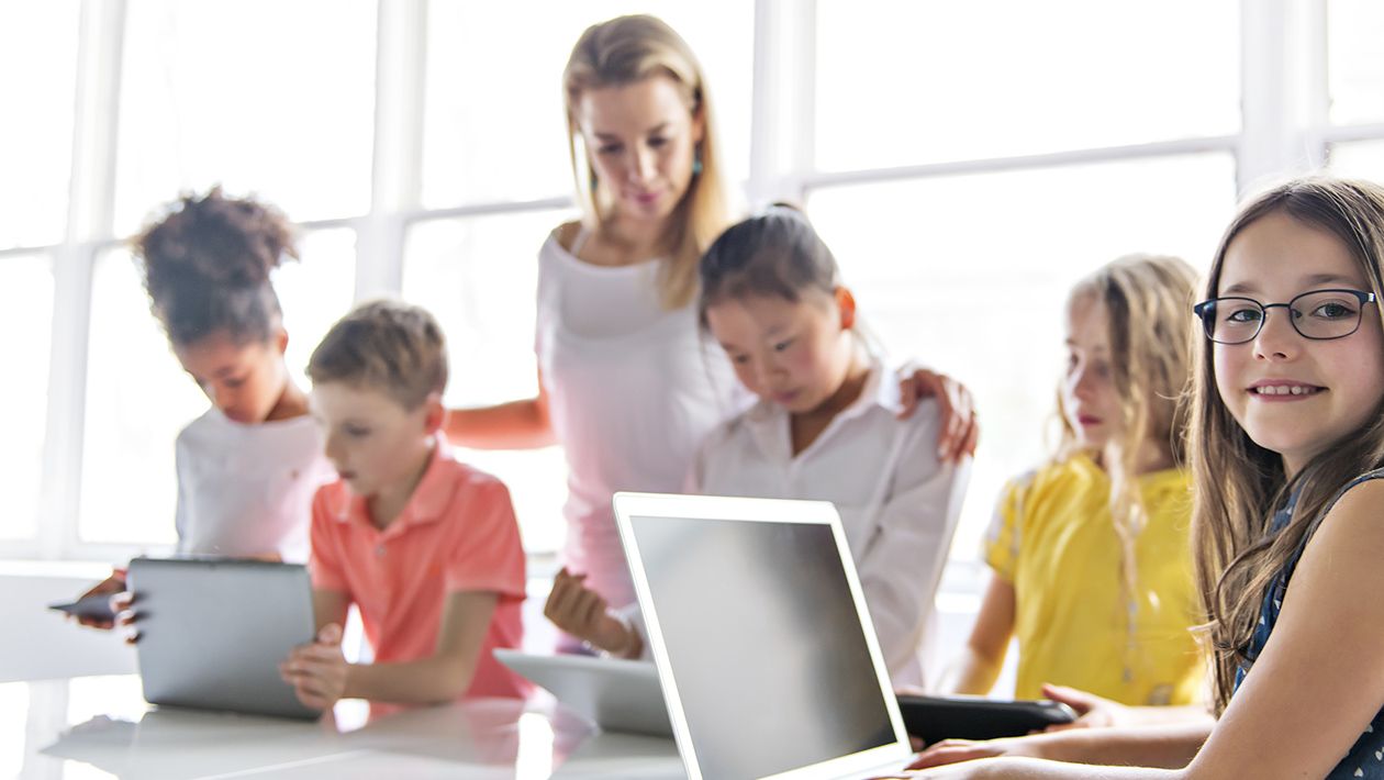 kids with tablets and laptop computer in classroom while teacher stands behind them
