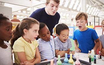 Teacher and kids watch experiment where they add colorful liquid into beakers